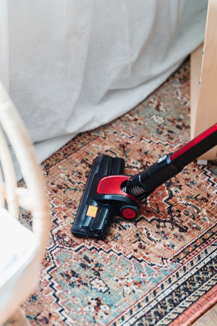 Close-up of a handheld vacuum cleaner with a red and black body performing surface cleaning on a traditional patterned area rug, situated in a room with a light-colored curtain and wooden furniture. The rug exhibits intricate designs and a rich color palette, and appears clean and well-maintained. The lighting is natural and soft, highlighting the textures of the fibers. This scene reflects a deep cleaning process for inherited rugs, as part of domestic cleaning services provided by Marylebone Carpet Cleaners, focusing on hygienic maintenance and surface sanitisation.