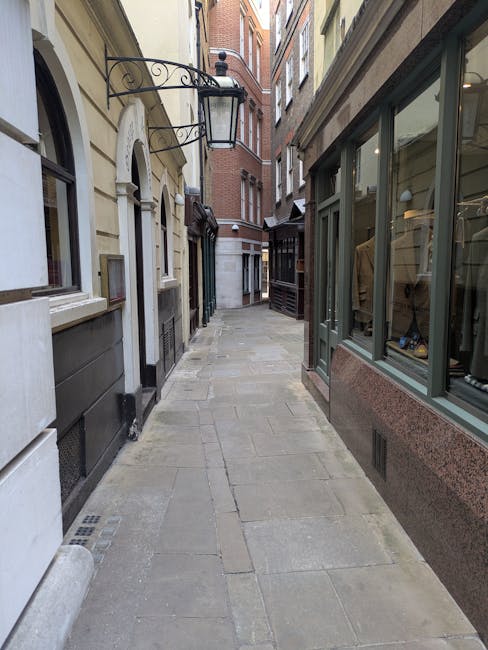View from inside a boutique on Chiltern Street, showing a bright blue wooden door slightly ajar, leading to the street where parked cars and a yellow vehicle are visible. The interior features a black-and-white checkered tile floor, a tall white shelving unit stocked with wine bottles, a small wooden chair with a green cushion, and a plush, round, teal seat. The wall above the shelf has floral patterned wallpaper, and natural light filters through the open door, illuminating the space. The setup suggests a clean, well-maintained environment, highlighting the importance of surface cleaning and maintenance, consistent with services provided by Marylebone Carpet Cleaners, to ensure hygienic and attractive interior and exterior surfaces.