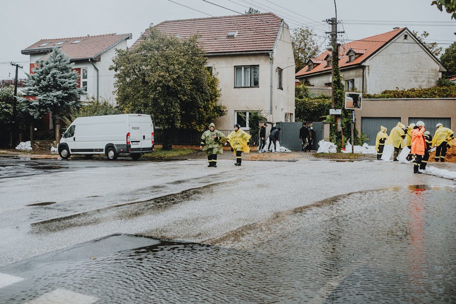 A residential street scene during a flood response, showing a wet, reflective asphalt surface with pooled water and tire tracks. Several workers in yellow and green raincoats and protective gear are actively engaged in flood cleanup efforts near a row of houses with white and cream-colored exteriors, tiled roofs, and various trees and shrubs in the background. A white service van is parked on the side of the street, and tools or equipment are not distinctly visible. The overcast sky and rain-soaked environment highlight the ongoing emergency basement flood cleanup coordinated by Marylebone Carpet Cleaners as part of their cleaning services.