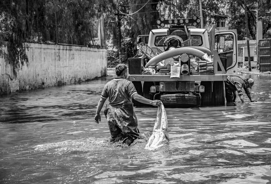 Black and white image depicting a worker in a rain-soaked area, holding a cloth or bag, with water partially submerging the ground. In the background, a large industrial water pump with hoses attached is positioned on a flooded street or driveway. The surfaces include wet pavement and concrete walls, reflecting the wet conditions, and the scene suggests a recent or ongoing flood cleanup process. The lighting highlights the reflective wet surfaces, emphasizing the severity of the flooding. The presence of the equipment and the worker's attire indicates a professional effort in surface cleaning and flood decontamination. Marylebone Carpet Cleaners offers expert flood damage restoration and deep cleaning services, ensuring thorough sanitisation of affected areas.