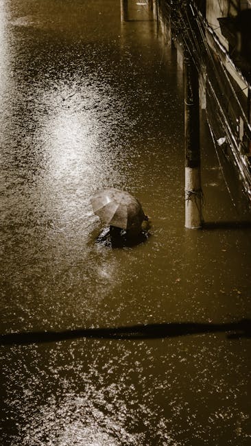 A residential street scene during a flood response, showing a wet, reflective asphalt surface with pooled water and tire tracks. Several workers in yellow and green raincoats and protective gear are actively engaged in flood cleanup efforts near a row of houses with white and cream-colored exteriors, tiled roofs, and various trees and shrubs in the background. A white service van is parked on the side of the street, and tools or equipment are not distinctly visible. The overcast sky and rain-soaked environment highlight the ongoing emergency basement flood cleanup coordinated by Marylebone Carpet Cleaners as part of their cleaning services.