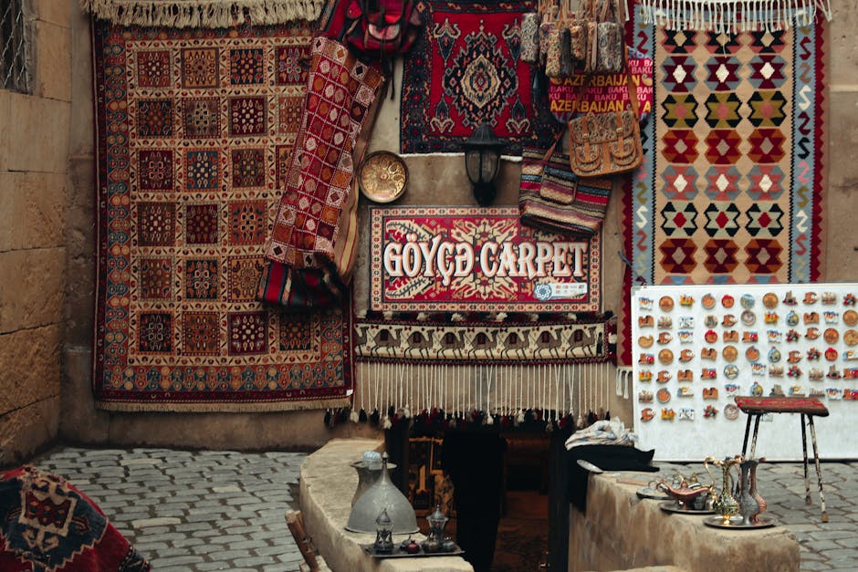 A display of intricately patterned Persian and Oriental rugs hanging against a stone wall at an outdoor market stall. The rugs feature vibrant geometric and floral designs in red, blue, gold, and cream hues. Below the rugs, a white table showcases small decorative items such as pottery, jewelry, and souvenirs. The ground is cobblestone, and there is natural lighting illuminating the scene. The setting reflects a traditional market environment, with a focus on the vibrant, clean, and well-maintained surfaces, suitable for discussion of surface cleaning and maintenance as part of Marylebone Carpet Cleaners' services.