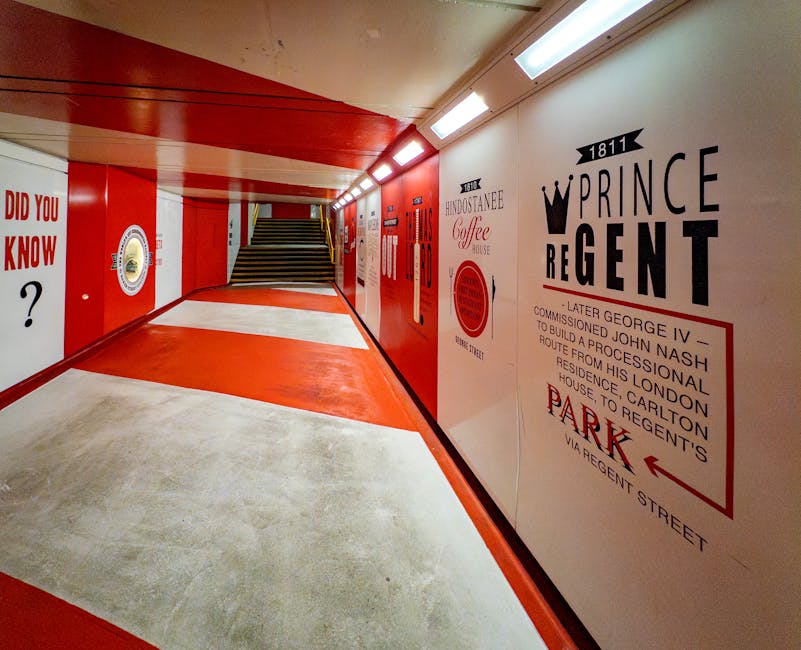 Interior view of a modern pedestrian walkway featuring a polished concrete floor with red and white painted sections, clean and well-maintained, leading to stairs. The walls are adorned with vintage-style advertising murals in black, red, and white, mentioning historic locations and brands such as Prince Regent and Hindostanee Coffee House. The ceiling is fitted with bright, linear LED lights, illuminating the space evenly. The area is free of dust and debris, with a sleek, hygienic appearance, reflecting thorough surface cleaning and maintenance by Marylebone Carpet Cleaners to ensure a hygienic environment for visitors.