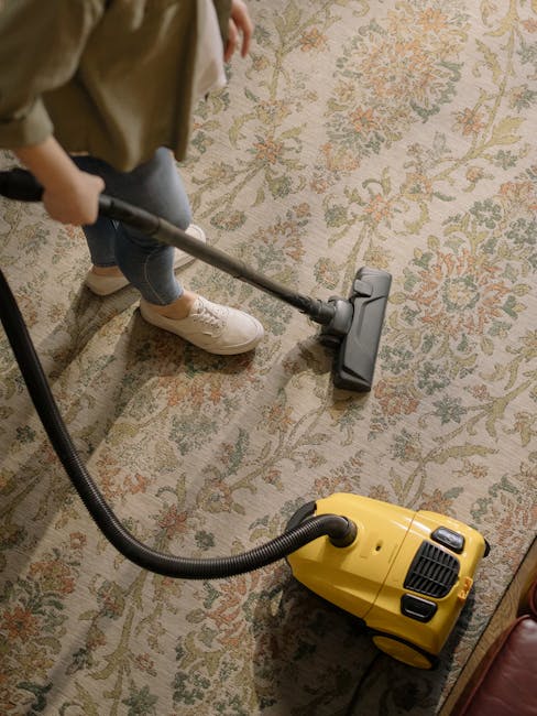 A person is engaged in surface cleaning using a vacuum cleaner equipped with a hose, focused on a patterned, traditional-style carpet in a residential room. The carpet features a floral design with muted tones of green, beige, and orange. The individual is wearing beige shoes, blue jeans, and a light-colored jacket. The vacuum cleaner, which is yellow with black accents, is positioned on the carpet, with the hose extended towards the area being cleaned. The room is well-lit, highlighting the cleanliness and detailed pattern of the carpet, and the scene suggests a professional deep cleaning process carried out by Marylebone Carpet Cleaners for effective sanitisation and odour removal in a domestic setting.