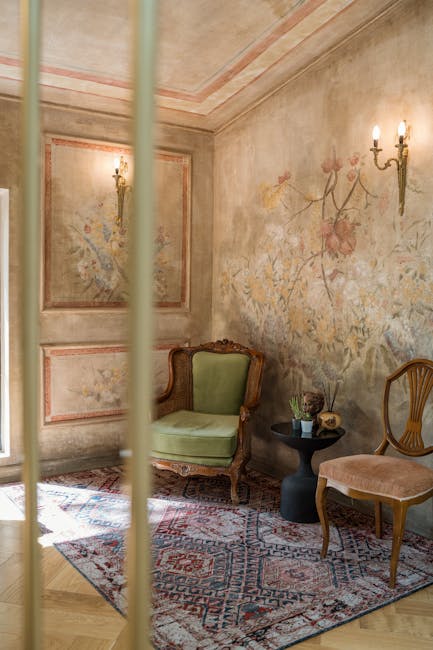 A corner of a room in the Portman Square terraces featuring antique furnishings and decor. The space has ornate pastel-colored walls with a floral mural and decorative molding, illuminated by warm light from a vintage wall sconce with two candles. There is a green upholstered armchair with wooden carved arms, a small round black side table holding decorative items, and a wooden chair with a curved back and cushioned seat. The room's wooden flooring is partially covered by an intricately patterned oriental rug, and the overall setting appears well-maintained and clean, reflecting the expertise of Marylebone Carpet Cleaners in surface cleaning and deep cleaning for historic interiors.