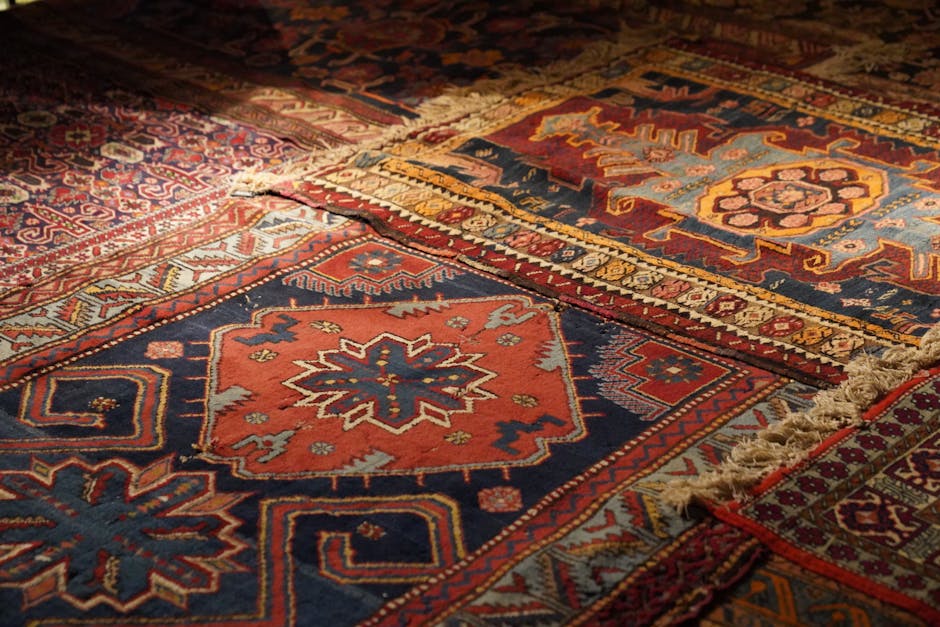Close-up view of multiple antique area rugs with intricate geometric patterns and rich colors, including reds, blues, and golds, laid out on a wooden floor in a well-lit room. The rugs display detailed woven designs with some fringed edges visible. The surfaces appear clean and well-maintained, reflecting professional deep cleaning and surface sanitisation by Marylebone Carpet Cleaners, suitable for preserving and caring for valuable, historical textiles in residential settings.