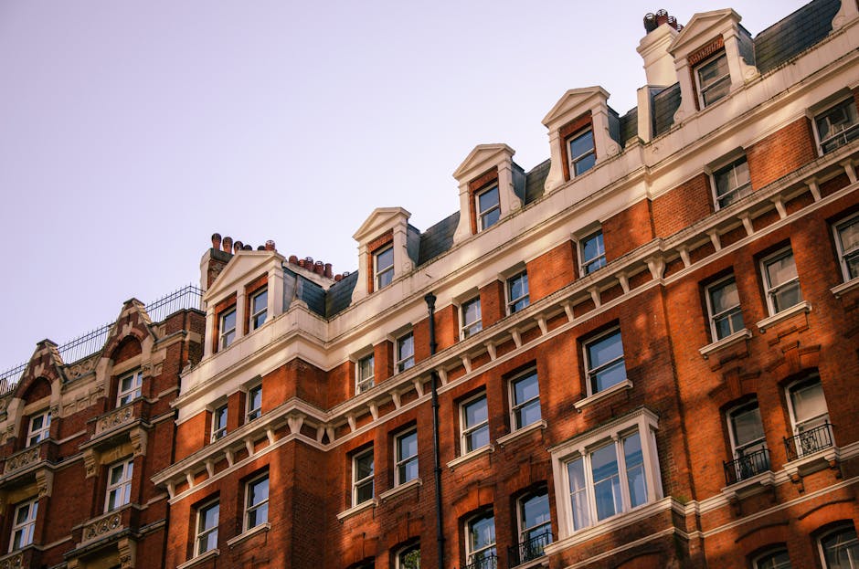 The image displays the upper floors of a traditional red brick residential building located in Marylebone, featuring multiple windows with white trim, some of which are open. The building has a classic Victorian architectural style with ornate white cornices and decorative elements along the roofline. The windows reflect blue sky, indicating clear weather, and the surrounding environment appears bright with natural sunlight casting soft shadows. The rooftop includes chimney stacks and a small black rainwater downpipe running down the side. The building's facade looks well-maintained, clean, and free of any visible mould or dirt, suggesting regular upkeep. Visible elements include window frames, glass panes, and brickwork, emphasizing surface cleanliness and architectural detail, which aligns with the expertise of Marylebone Carpet Cleaners in surface cleaning and hygiene maintenance.