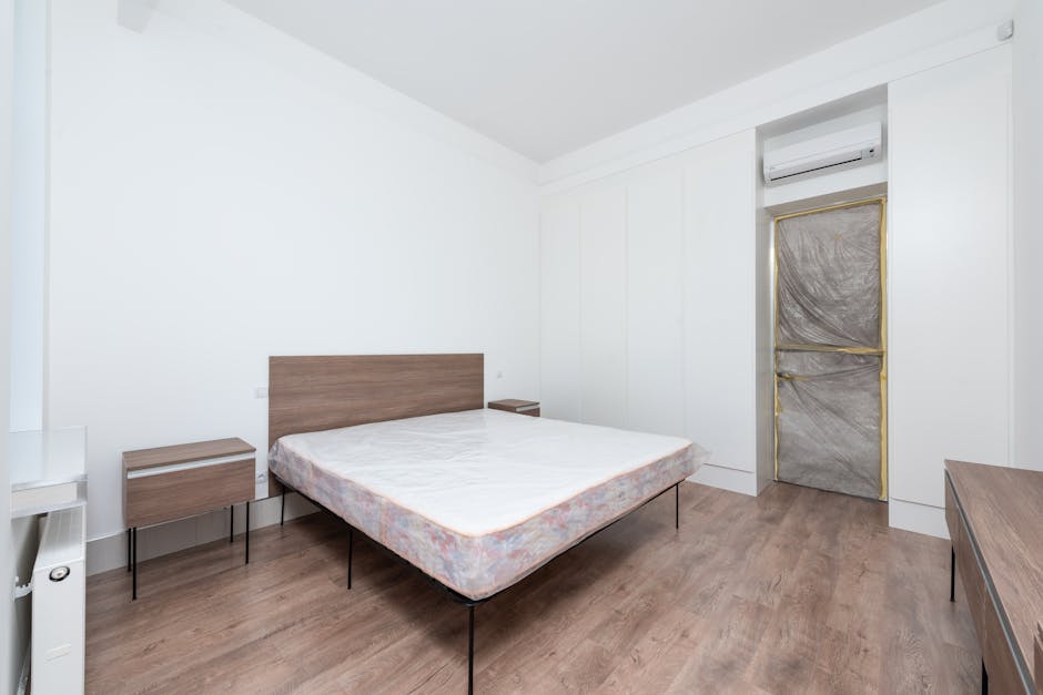 A minimalist bedroom featuring a double bed with a wooden headboard and a clean, unmade mattress. Adjacent to the bed are two small bedside tables made of matching wood with black metal legs. The room has light-colored walls and wooden flooring, with a white ceiling. On the right side, there's a door covered with protective brown paper and secured with tape, likely for renovation or maintenance purposes. Above the door, an air conditioning unit is mounted on the wall. The room is well-lit with natural and artificial light, emphasizing the cleanliness and tidiness of the space, which reflects professional surface cleaning and maintenance typical in residential environments managed by Marylebone Carpet Cleaners for hygiene and mould prevention purposes.
