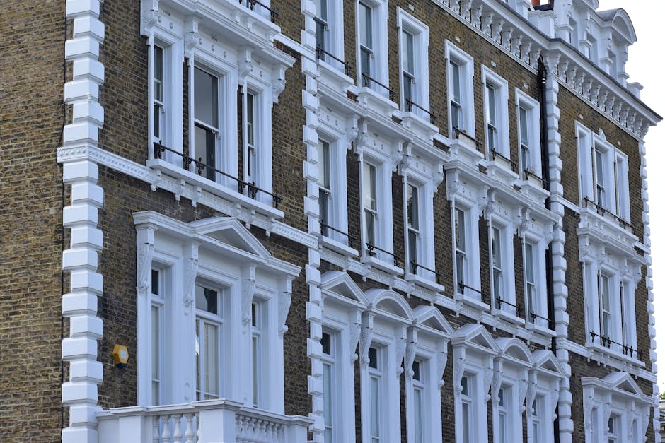The image displays the upper floors of a traditional red brick residential building located in Marylebone, featuring multiple windows with white trim, some of which are open. The building has a classic Victorian architectural style with ornate white cornices and decorative elements along the roofline. The windows reflect blue sky, indicating clear weather, and the surrounding environment appears bright with natural sunlight casting soft shadows. The rooftop includes chimney stacks and a small black rainwater downpipe running down the side. The building's facade looks well-maintained, clean, and free of any visible mould or dirt, suggesting regular upkeep. Visible elements include window frames, glass panes, and brickwork, emphasizing surface cleanliness and architectural detail, which aligns with the expertise of Marylebone Carpet Cleaners in surface cleaning and hygiene maintenance.