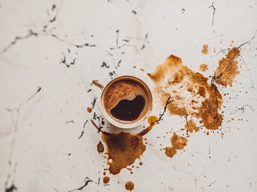 A top-down view of a white surface with visible cracks and black markings, featuring a coffee mug filled with dark coffee and frothy bubbles on top. Surrounding the mug are several large coffee stains with irregular shapes and splatters, indicating recent spills. The surface appears to be a desk or countertop with a matte finish, and the stains contrast strongly against the white background. Visible in the image are parts of the coffee mug, which is situated centrally, and the stains seem fresh, possibly from recent accidental spills. The scene highlights the importance of surface cleaning and stain removal, as offered by Marylebone Carpet Cleaners, known for their deep cleaning and sanitisation services in Marylebone.